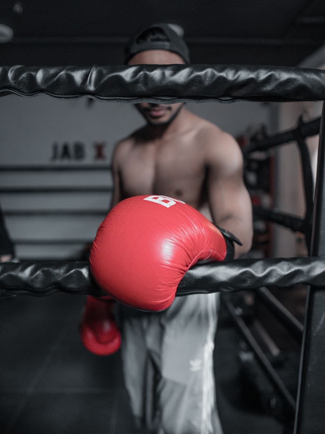Shirtless man with face obscured by rope resting one large red glove on rope below