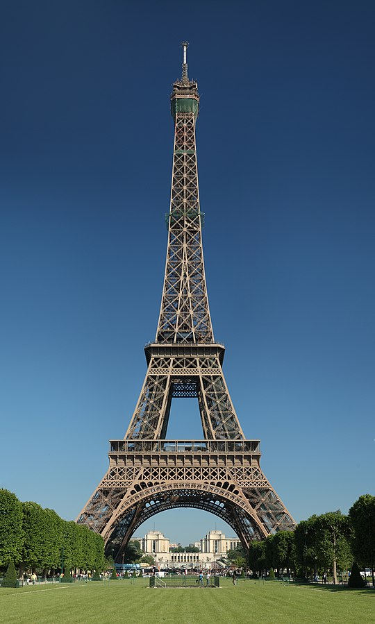The Eiffel Tower with its iconic lattice design against a blue background surrounded by greenery and people