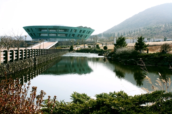 Color photograph shot across a long, clear body of water with a green structure and slope in the distance