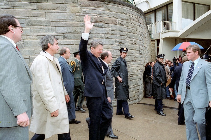 Black and white photo of Ronald Reagan waving to the crowd surrounded by men in suits shortly before assassination attempt